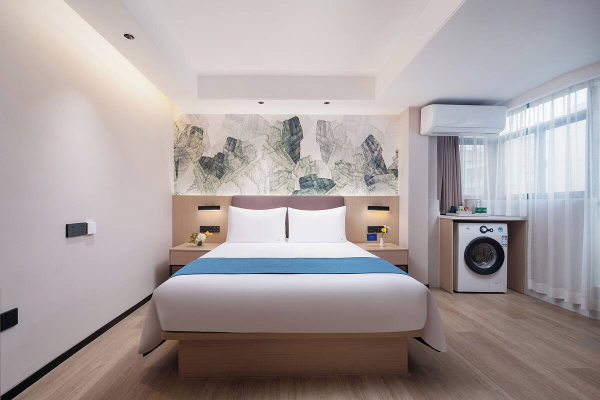 A modern hotel room with a large bed, a washing machine, and a view of the city through large windows.