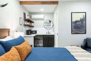 Quarto com cama, almofadas azuis e laranjas, uma pequena cozinha, um espelho e um cartaz de bicicleta.