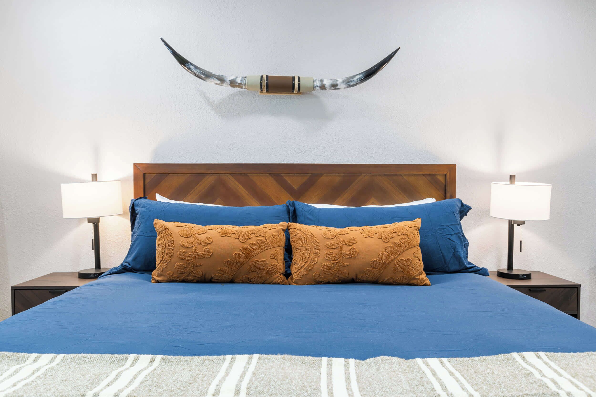 A bed with a blue bedspread and two brown pillows, flanked by two bedside tables with lamps, against a white wall with a mounted horn decoration.