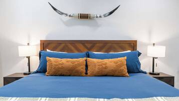 A bed with a blue bedspread and two brown pillows, flanked by two bedside tables with lamps, against a white wall with a mounted horn decoration.