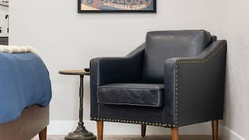 Una habitación con un sillón de cuero oscuro, una mesita redonda, una alfombra estampada y una cama con cubrecama azul.