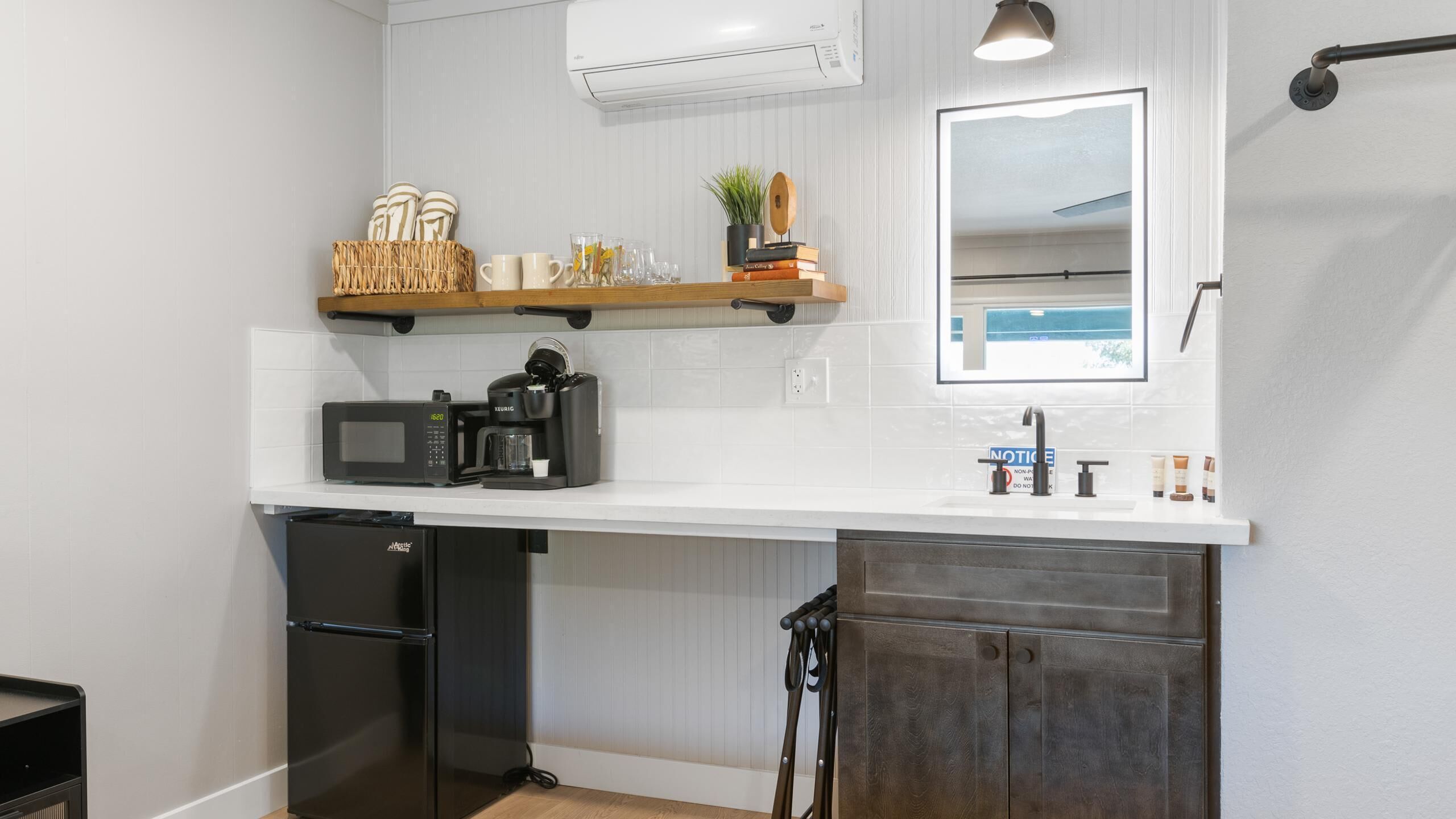 A compact kitchen with a microwave, coffee maker, and a small refrigerator. There is a wooden shelf above the counter holding books and a plant. A mirror is mounted on the wall above the counter.
