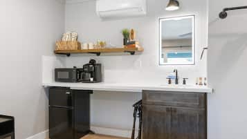 A compact kitchen with a microwave, coffee maker, and a small refrigerator. There is a wooden shelf above the counter holding books and a plant. A mirror is mounted on the wall above the counter.
