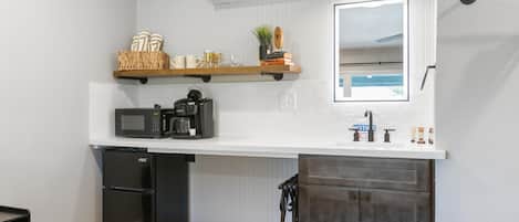 A compact kitchen with a microwave, coffee maker, and a small refrigerator. There is a wooden shelf above the counter holding books and a plant. A mirror is mounted on the wall above the counter.