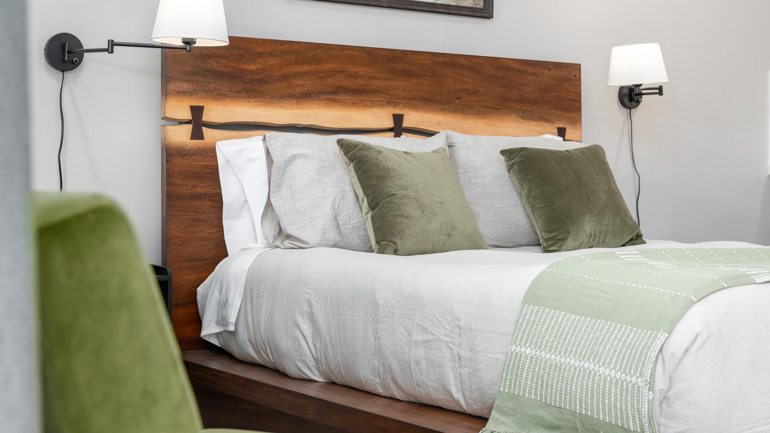 Un lit bien fait, avec une tête de lit en bois, une literie blanche et des coussins verts. Deux lampes murales sont fixées de chaque côté du lit.