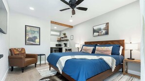 Un dormitorio con cama, una silla, ventilador de techo, lámpara y un cuadro en la pared.