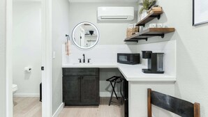 A compact kitchen with a white countertop, a black microwave, and a sink. There are wooden shelves above the counter and a small stool. - Sequoia Motel King RM6, AC, Pool, WiFi 10 Min SNP (Three Rivers)