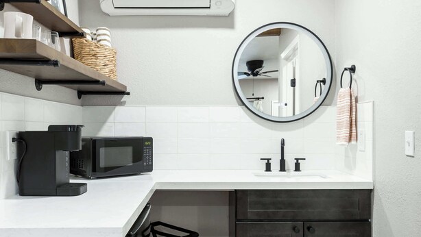 A modern bathroom with a white countertop, a black coffee machine, a microwave, a sink with black faucets, a round mirror, and a ceiling fan. - The Rae Sequoia Motel RM5 (Three Rivers)