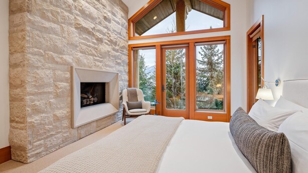 A bedroom with a stone fireplace, a bed, a chair, and large windows with a view of trees. - Alpine chalet w/ AC, hot tub, media (Whistler)
