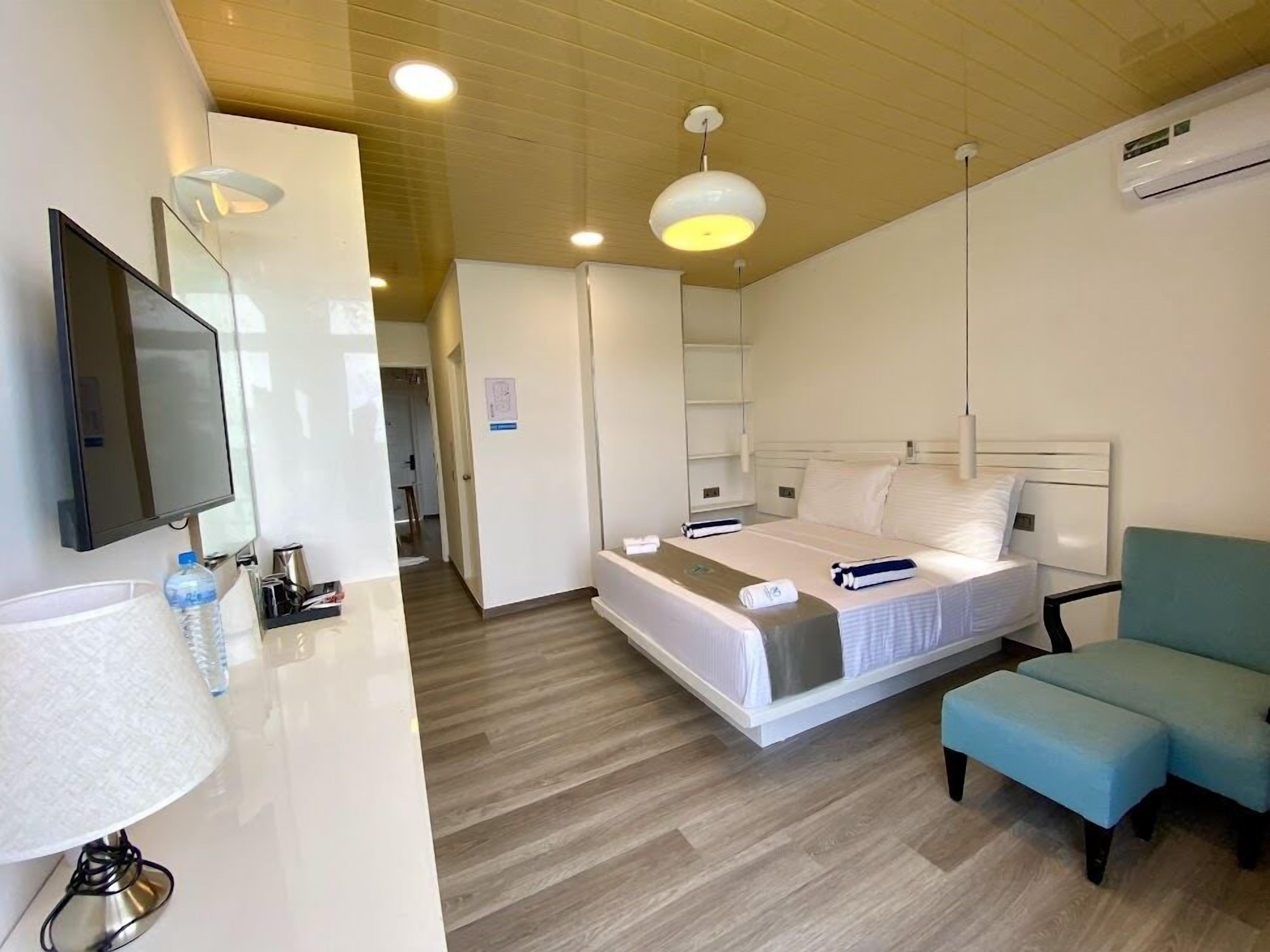 Una habitación de hotel moderna con una cama, una mesita de noche con lámpara, un televisor y un rincón de estar con un sillón.