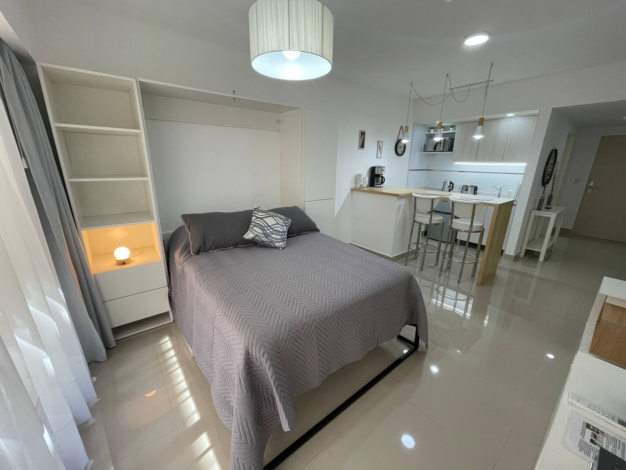 Ein modernes Schlafzimmer mit Bett, Nachttisch, integriertem Kleiderschrank und einer kleinen Küchenzeile im Hintergrund.