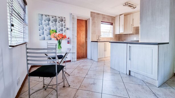 A modern kitchen with white cabinets, a dining area with a chair and table, and a decorative wall with apples. - Stylish one bed Apartment in Strydom Park - 2168 (Johannesburg)