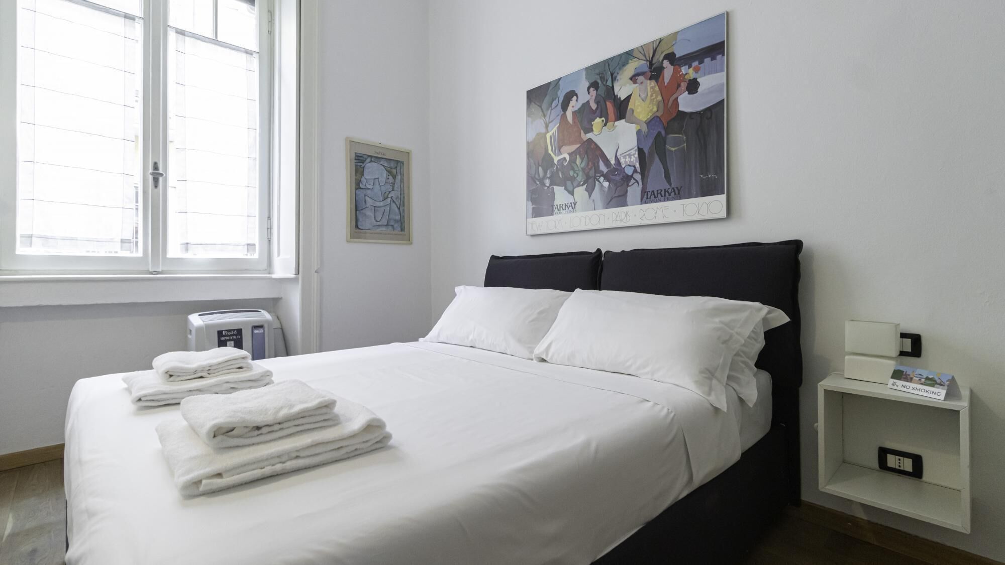 Ein Hotelzimmer mit Bett, Nachttisch, Wandbild und Fenster mit Jalousien.