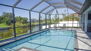 A covered outdoor swimming pool with clear blue water, surrounded by a paved deck and a view of a lake and greenery. - New Canal Access Pool Home w/Outdoor Oasis/Kayaks (Cape Coral)