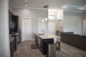 Una cocina moderna con una isla central, horno empotrado y refrigerador. Cuenta con un comedor con un taburete de bar y un sofá.