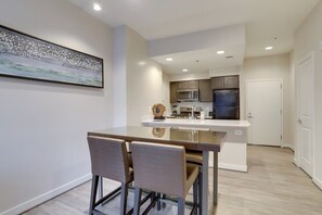A modern kitchen with stainless steel appliances, a dining area with a glass table and two chairs, and a white door in the background.
