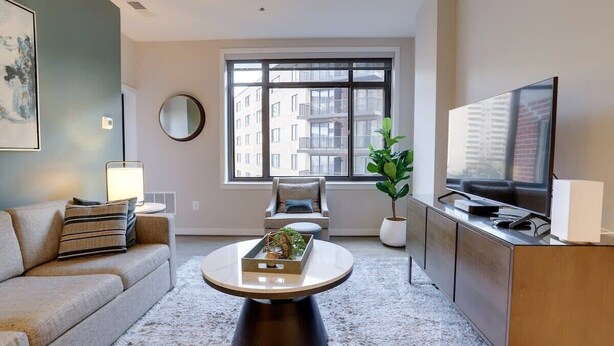 Een moderne woonkamer met een bank, salontafel, televisie en een groot raam met uitzicht op gebouwen.