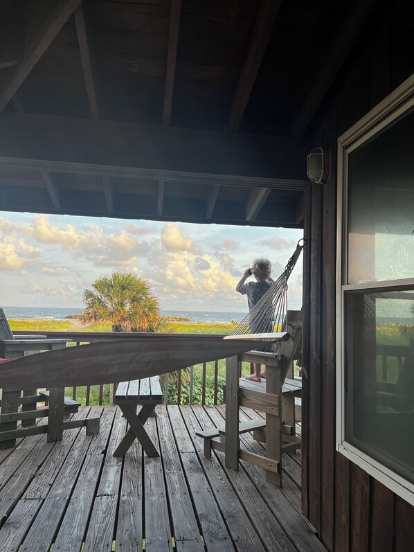 A wooden deck with a hammock, a table, and a view of the ocean and sky.