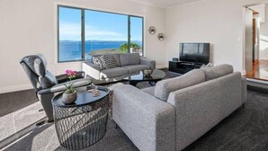 Una sala de estar moderna con un sofá gris, un sillón de cuero y una mesa de centro. Un televisor está montado en la pared y hay un ventanal con vistas al mar.