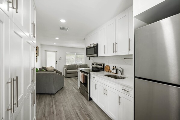 A modern kitchen with white cabinetry, stainless steel appliances, and a grey sofa in the adjacent living area. - PALM14 | delightful oasis in Pompano Beach (Pompano Beach)