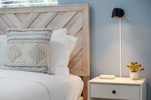 A neatly made bed with a wooden headboard, a white nightstand with a lamp, and a small potted plant. - PALM14 | delightful oasis in Pompano Beach (Pompano Beach)