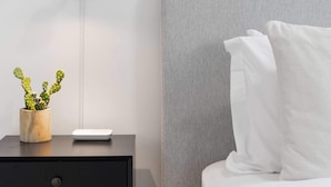 Un dormitorio moderno con cabecera gris, cama blanca, mesita de noche negra con lámpara y una pequeña planta de cactus.