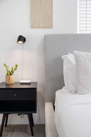 A modern bedroom with a grey headboard, a white bed, a black nightstand with a lamp, and a small cactus plant. - PALM14 | delightful oasis in Pompano Beach (Pompano Beach)