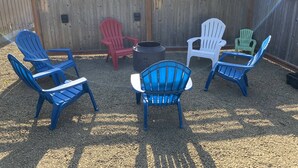 A fenced outdoor seating area with four Adirondack chairs, one red and three blue, arranged around a central fire pit.