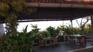 Un área de patio techada al aire libre con una mesa y sillas, rodeada de plantas en macetas y árboles.