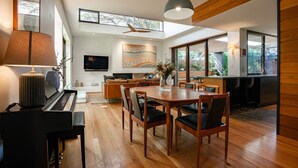 Een moderne woonkamer met een piano, eettafel en stoelen. Er is een televisie aan de muur bevestigd en een groot raam dat voor natuurlijk licht zorgt.