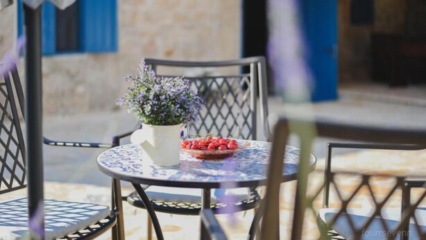 A round table with a floral centerpiece and a bowl of fruit, surrounded by metal chairs with a lattice backrest, set outdoors with a stone wall and blue shutters in the background. - Didini Dvori in Kru evo (Obrovac)