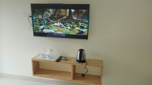 A television mounted on a wall displaying a nature scene. Below the TV, there is a wooden shelf with a kettle, glasses, and a remote control. - Hotel Anandam Phata (Ukhimath)