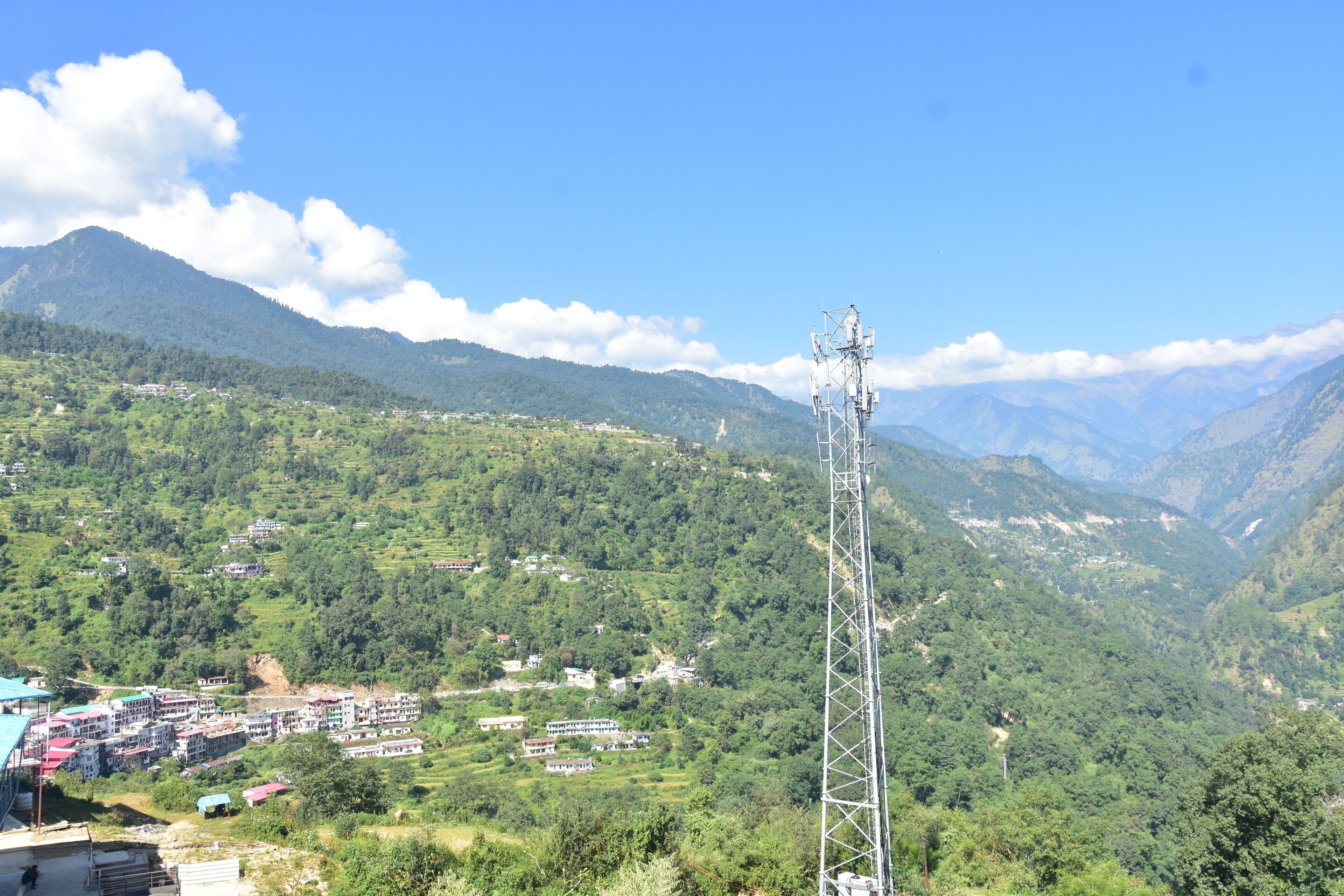 Un paisaje montañoso con una torre de telecomunicaciones, un pequeño asentamiento con edificios coloridos y colinas verdes exuberantes.