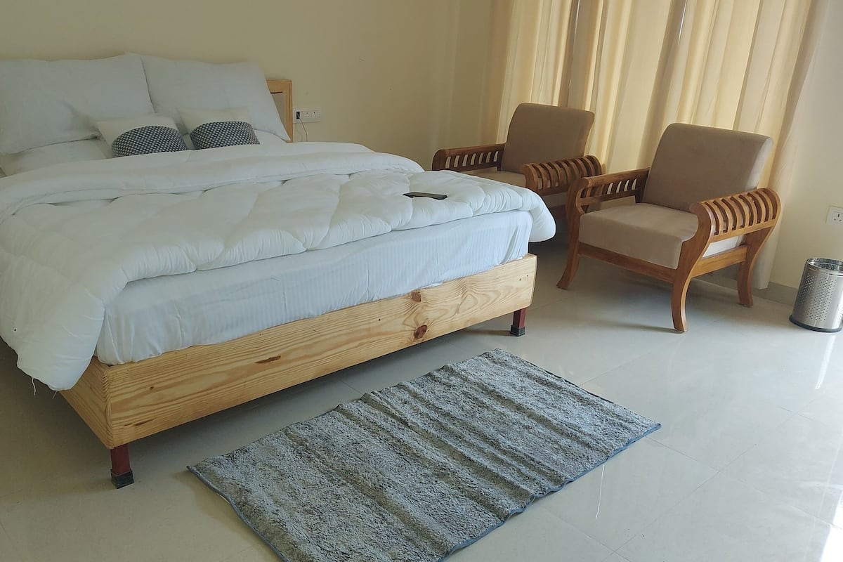A bedroom with a wooden bed, white bedding, two chairs, a small rug, and a trash can.