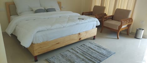 A bedroom with a wooden bed, white bedding, two chairs, a small rug, and a trash can.