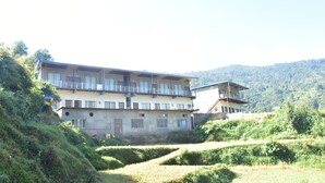 A multi-story building with a balcony on the upper floor, surrounded by terraced fields and a mountainous backdrop. - Hotel Anandam Phata (Ukhimath)