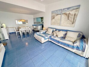 A modern living room with a blue tiled floor, a white sofa with patterned cushions, a dining area with white chairs, and a kitchen in the background.
