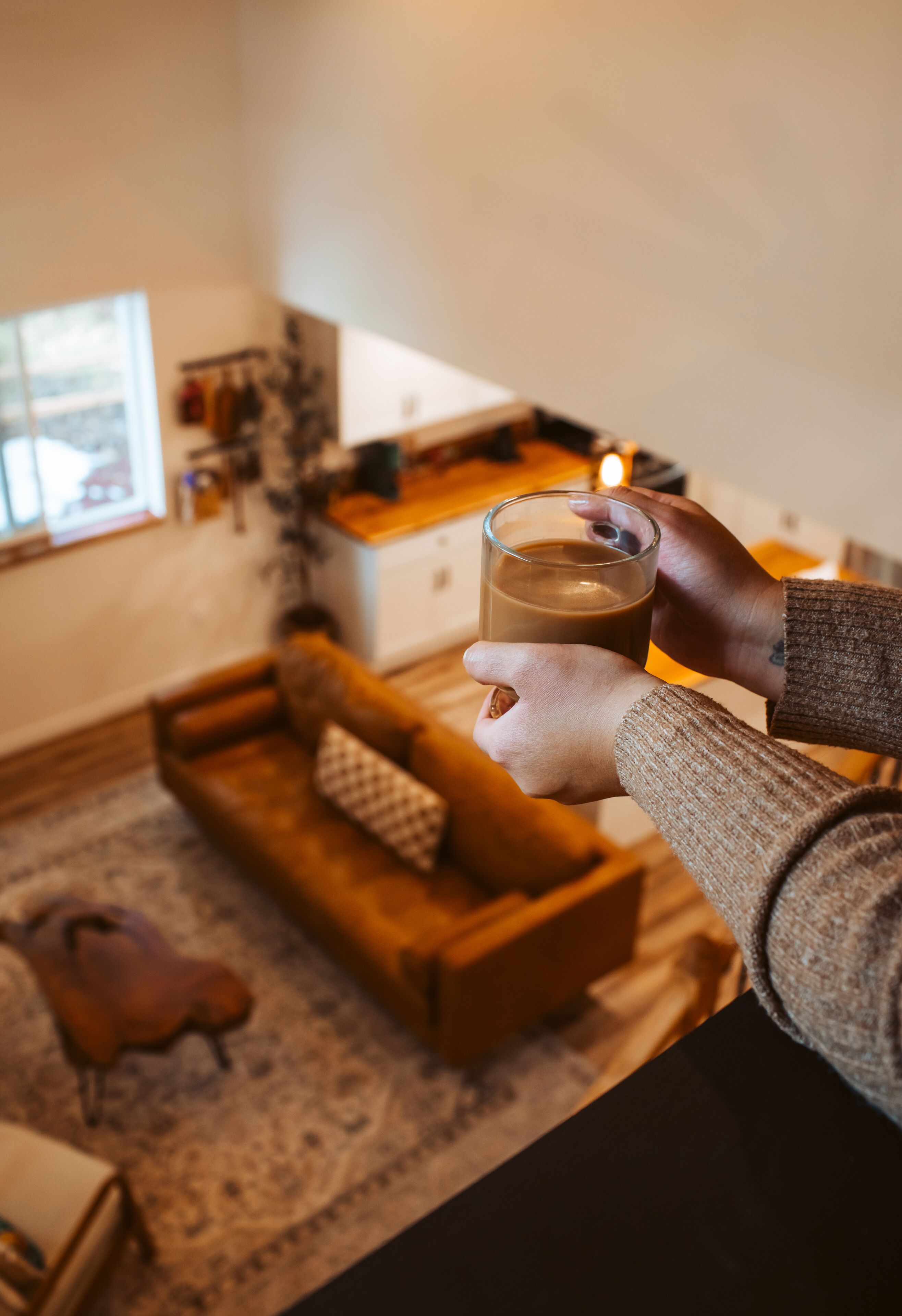 Uma pessoa segurando um copo de bebida em um quarto aconchegante, com um banco de madeira, uma mesinha e uma vela sobre a mesa.