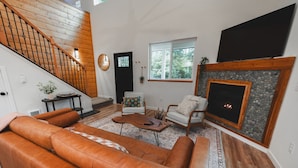 A living room with a brown leather sofa, a wooden staircase, a fireplace, and a large window.