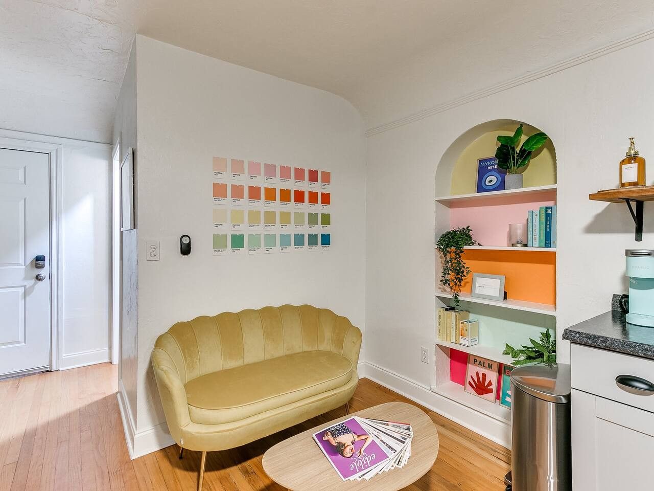 A room with a yellow sofa, a small round table with magazines, a bookshelf with various items, and a door.