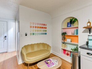 A room with a yellow sofa, a small round table with magazines, a bookshelf with various items, and a door.