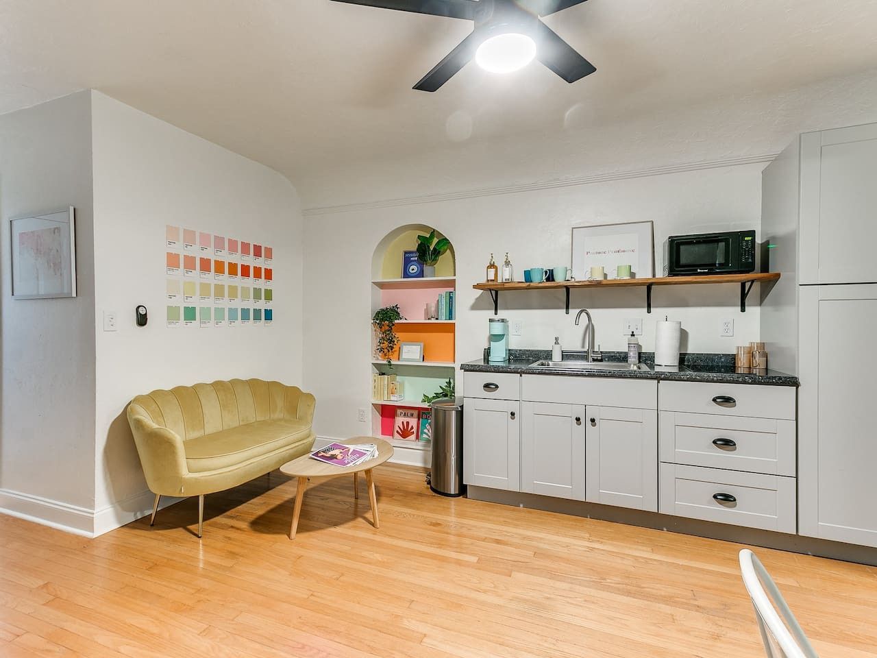 A modern kitchen with white cabinets, a microwave, and a coffee maker. There is a yellow sofa and a small table with a magazine.