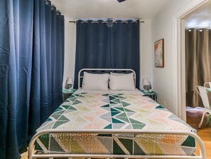 Ein Einzelbett mit gemustertem Bettbezug, ein Nachttisch mit Lampe und Vorhänge an beiden Seiten des Fensters.