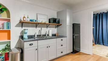 A compact kitchen with white cabinets, a microwave, and a refrigerator. There is a shelf with books and a potted plant.