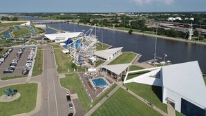 Eine Luftaufnahme eines Objets direkt am Wasser mit einem großen Parkplatz, einem Schwimmbecken und einem markanten, weißen Gebäude mit einem dreieckigen Dach.