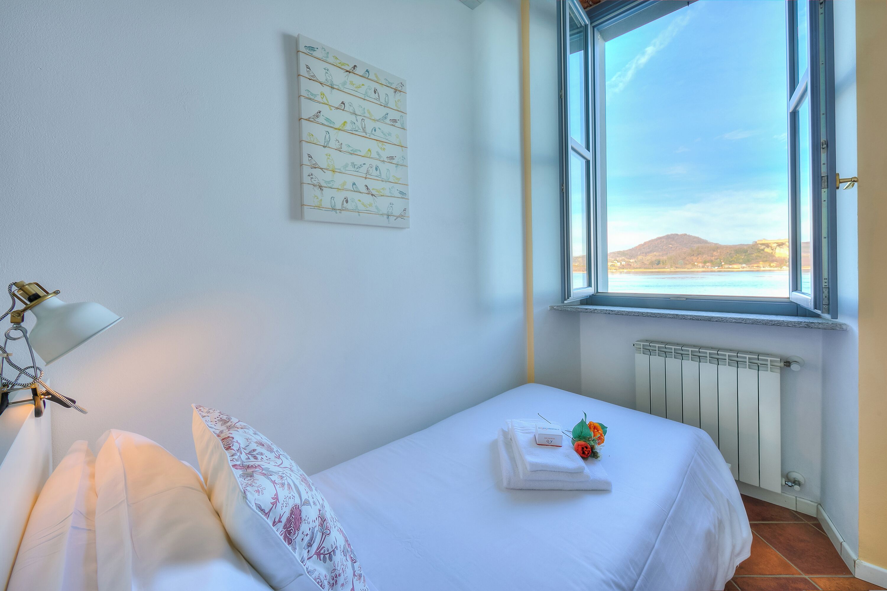 A bedroom with a bed, a window with a view of the sea, a radiator, and a wall with a bird illustration.