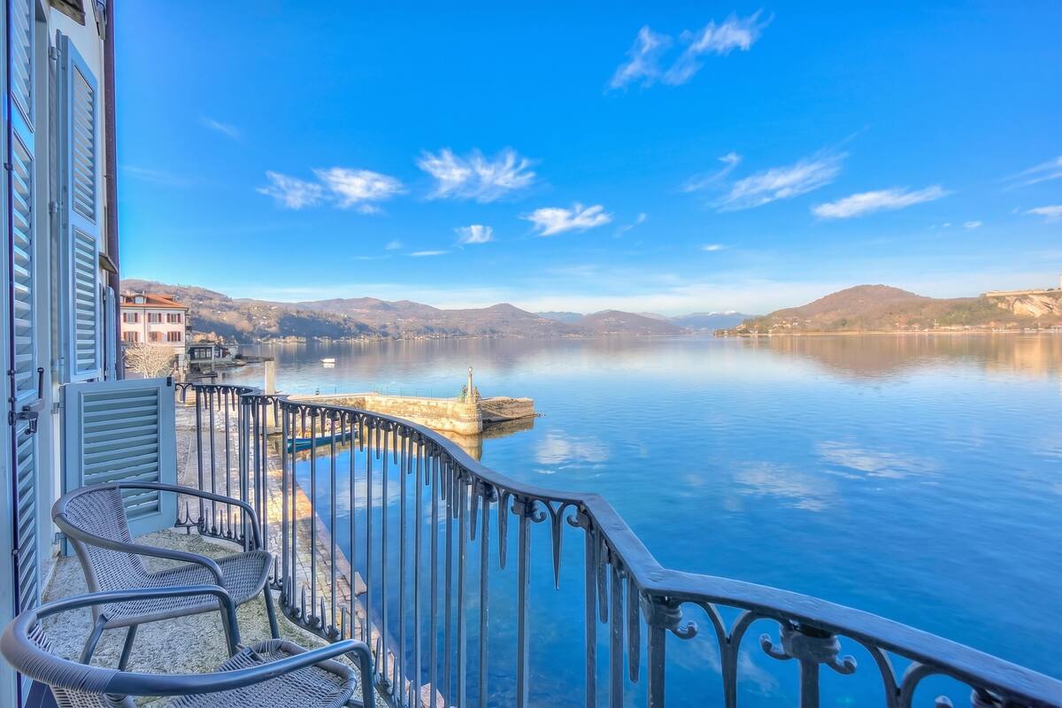 A balcony with a view of a calm lake, a chair, and a building with shutters.