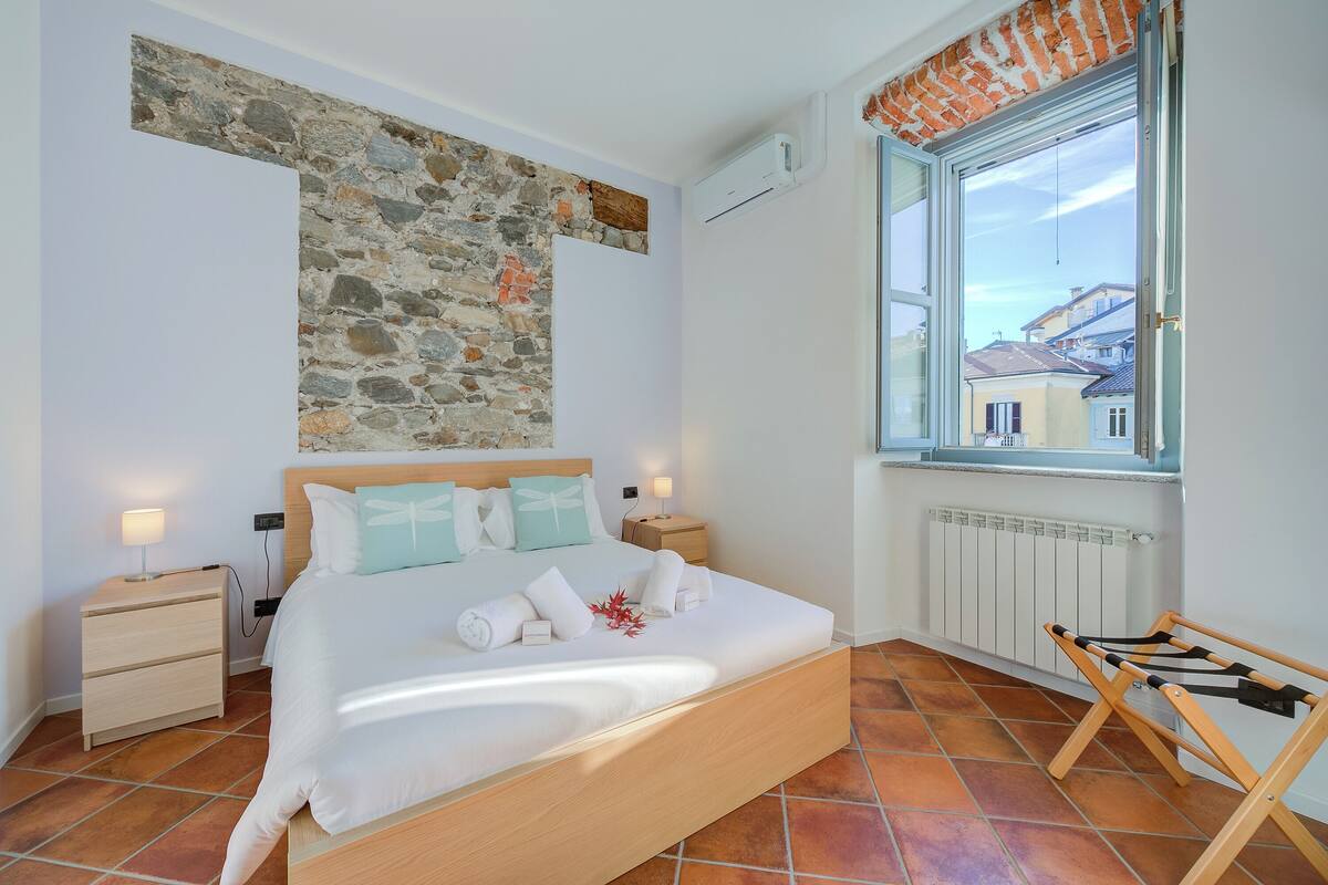 A bedroom with a stone wall, a bed with white bedding, a wooden nightstand, a window with a view of buildings, and a radiator.