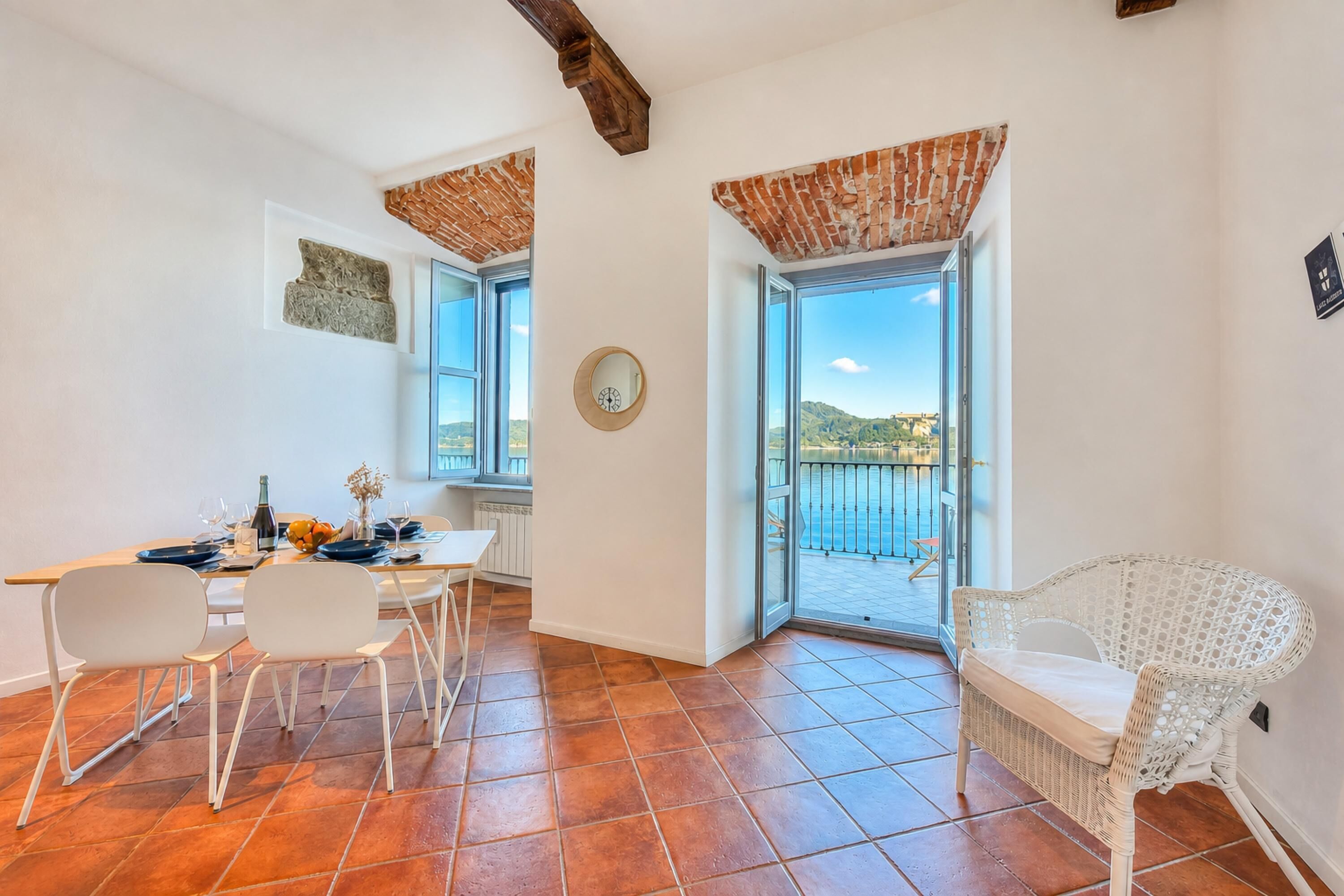 A dining area with a table set for two, a white wicker chair, and a view of the sea through a glass door.