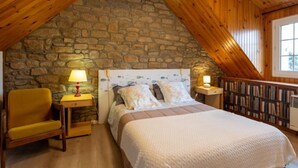 A cozy bedroom with a stone wall, wooden ceiling, a bed with a white quilt, a yellow chair, bedside lamps, and a bookshelf. - Esprit Bailly by Interhome (Cancale)